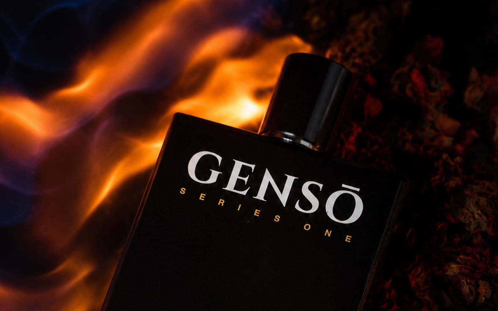 TAKUMI GENSŌ — the gloss-black bottle among dried rose petals with a wall of blue and orange flame rising behind it, cold and warm light held at once