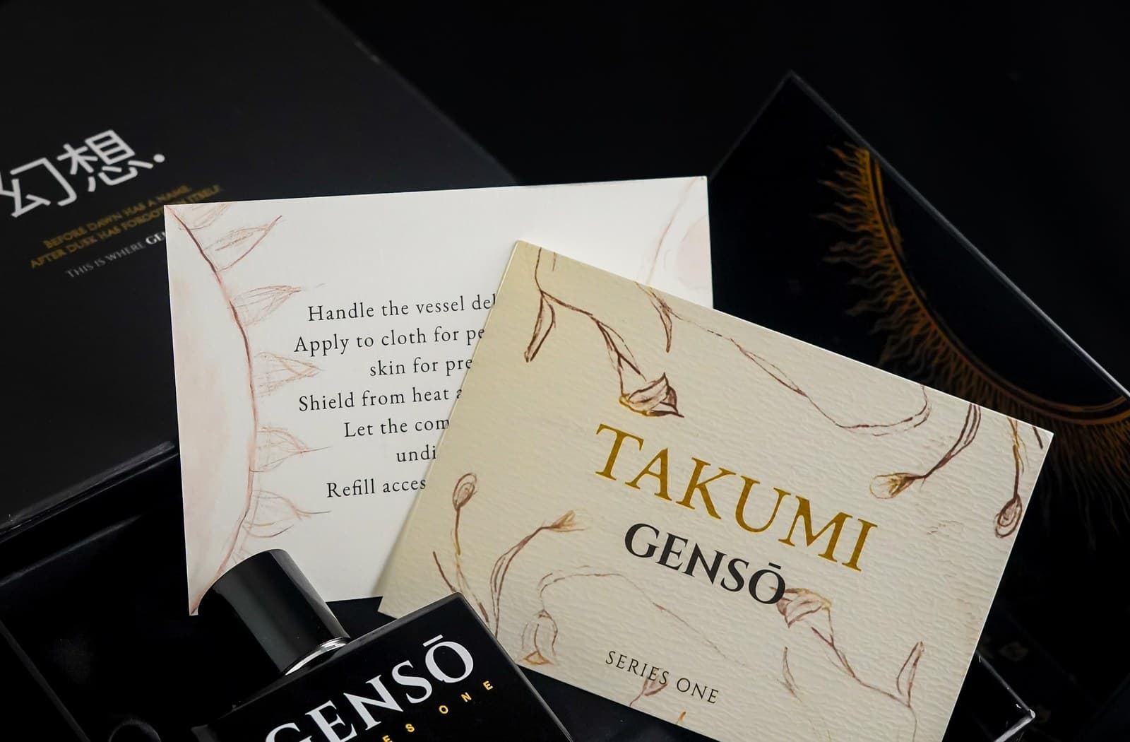 TAKUMI GENSŌ — the black box open, celestial sun-and-moon line work on the interior, the cream care card and TAKUMI card over the bottle in black satin