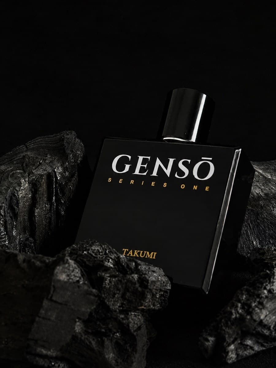 TAKUMI GENSŌ — gloss-black bottle upright in shadow, chrome atomizer exposed, cap resting beside it